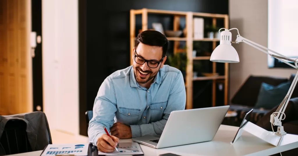 A man works at a white desk with papers, a laptop, and a lamp, in a modern, well-lit home office with shelves behind him.