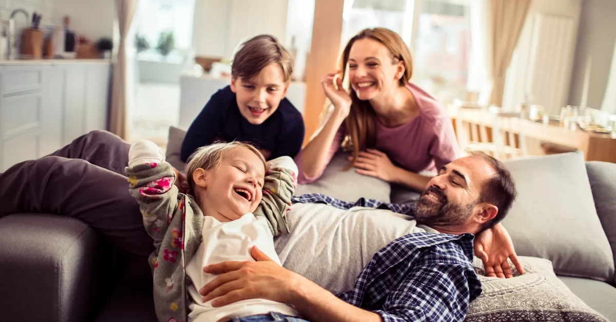 A joyful family of four plays on a light grey sofa in a bright, modern living room with a kitchen in the background.