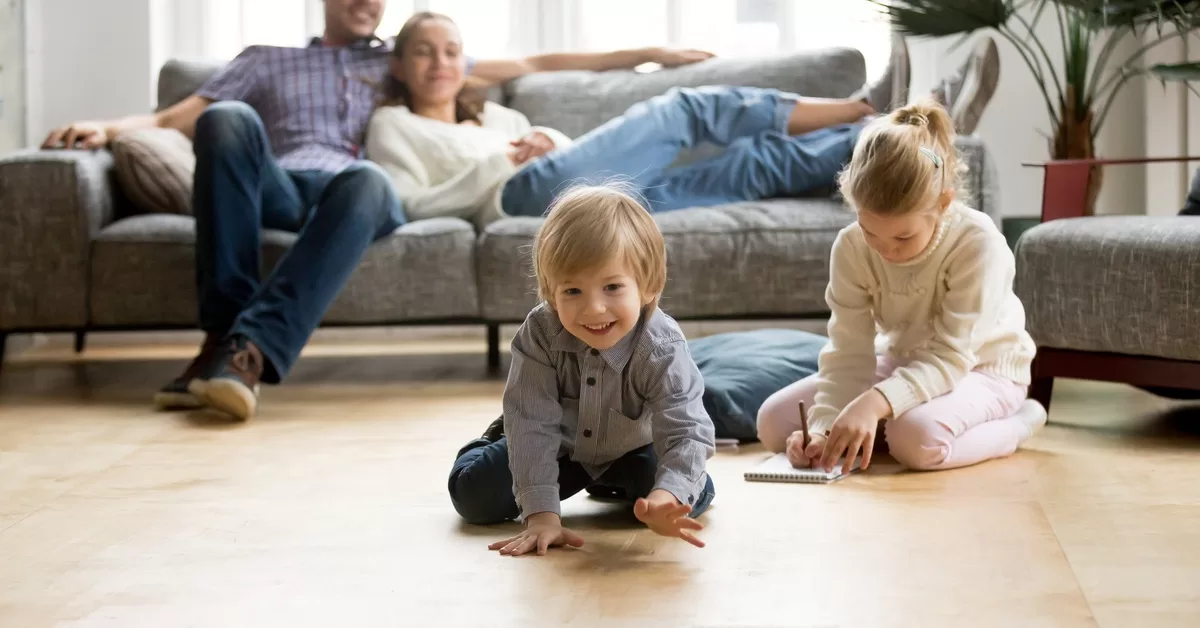 A family relaxes in a bright living room. The parents sit on a sofa while the kids play with a toy car and notebook.