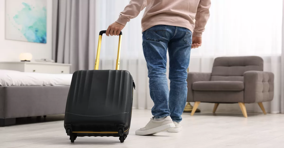 A person pulls a black suitcase with a golden handle in a well-lit room featuring a bed, armchair, and light curtains.