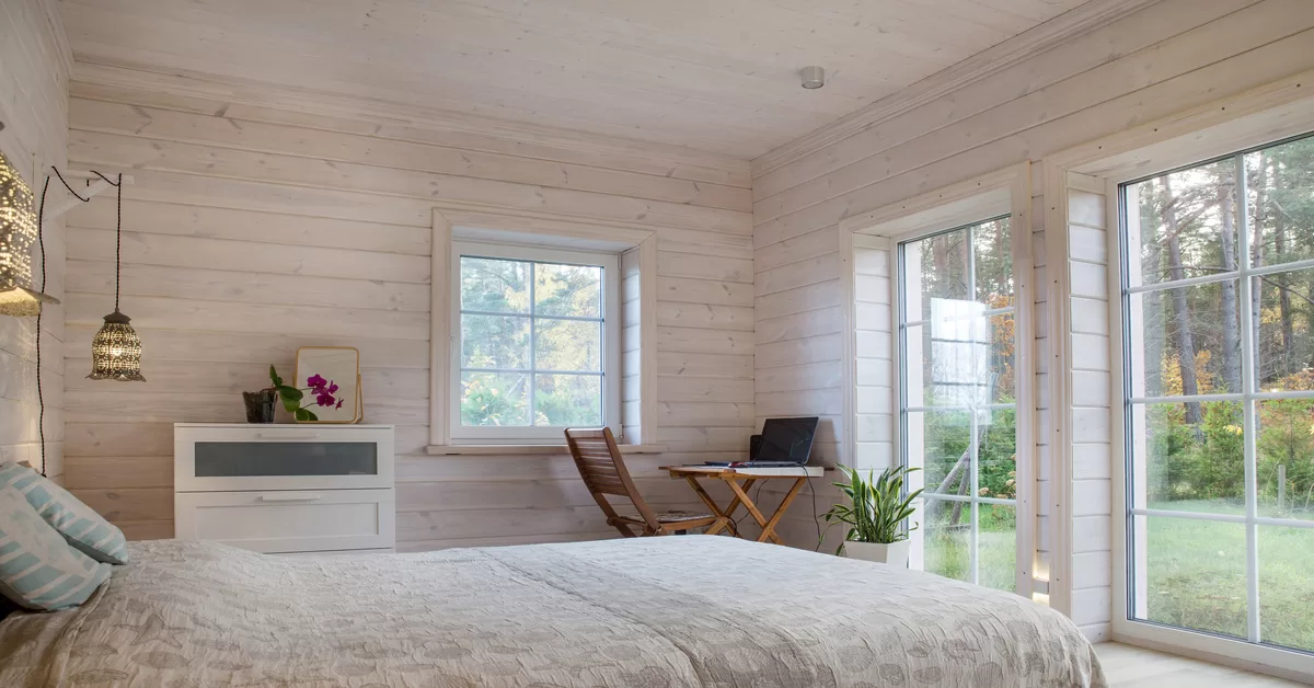 A bright bedroom contains wood paneling, a large bed, pendant lights, and floor-to-ceiling windows showing a forest view.
