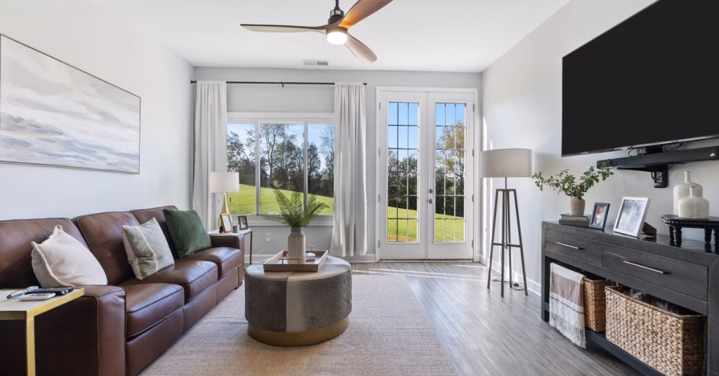 Bright modern living room with leather sofa, large windows, patio doors, wall-mounted TV, and views of a grassy landscape.