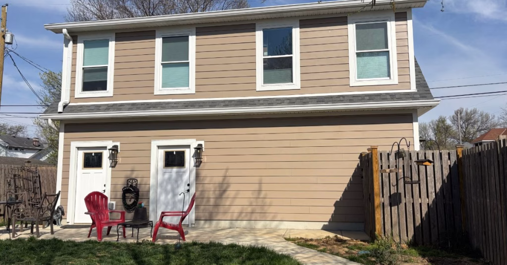 Rear view of a two-story garage apartment featuring beige siding, two entry doors, a small patio, and fenced backyard.
