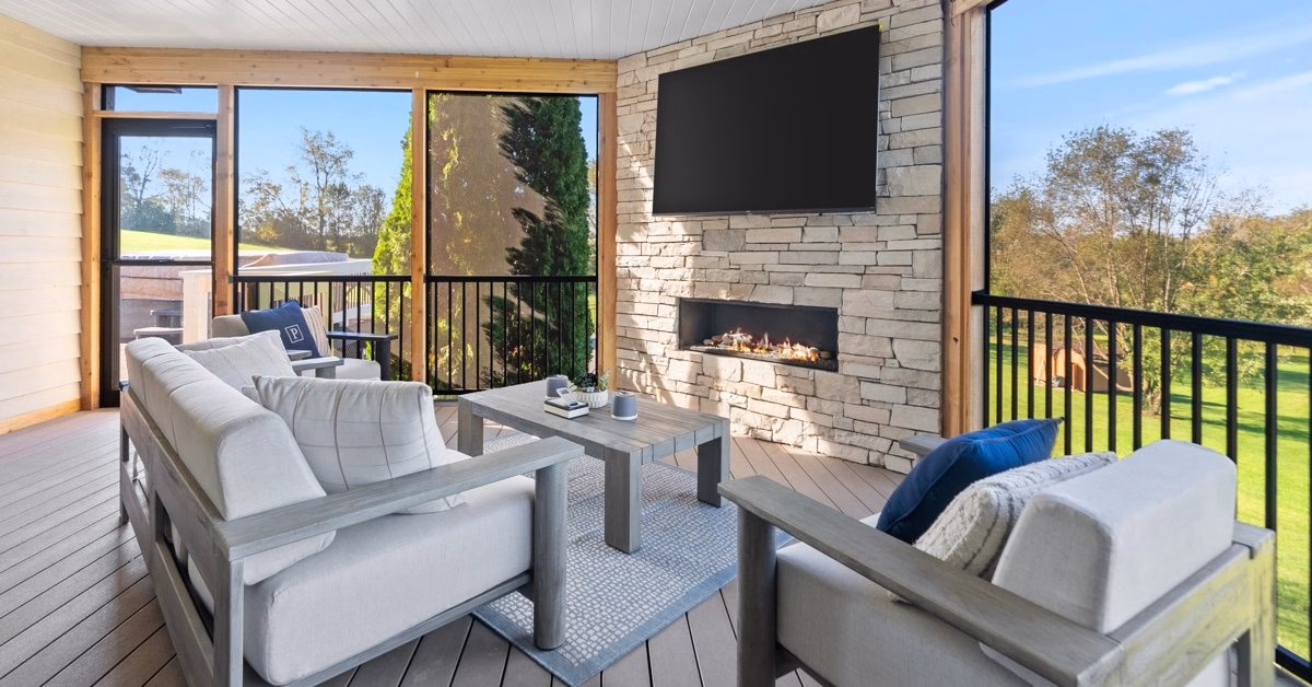 A covered outdoor living area with cushioned seating, a stone fireplace, and scenic views through screened windows.