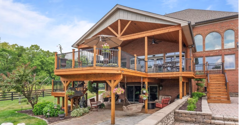 A covered wooden deck overlooks the patio and lawn behind a brick home with large windows and stairs.