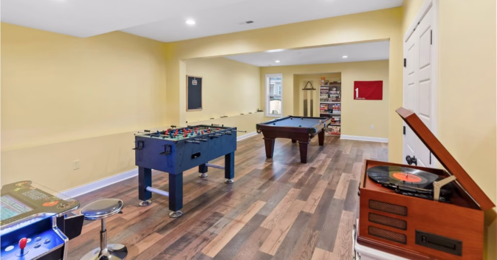 A finished basement game room includes a foosball table, a pool table, an arcade game, and warm wood flooring.