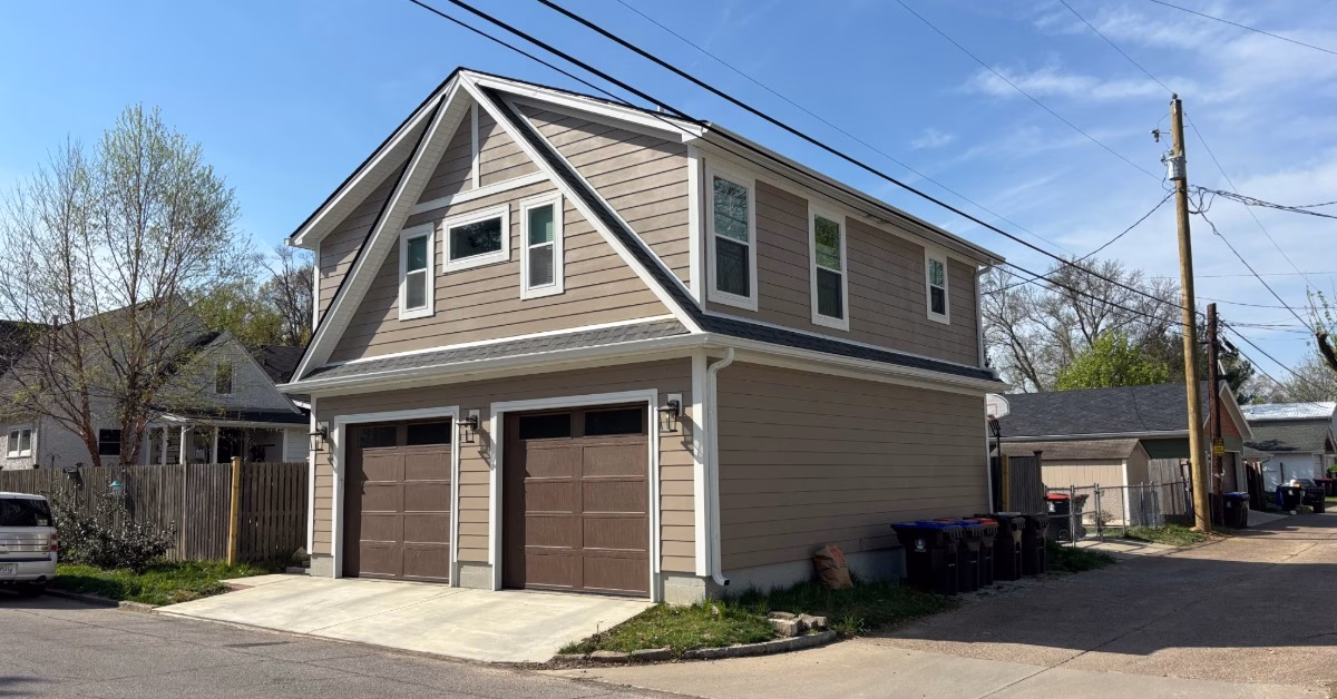 A tan two-story garage with brown double doors, white trim, overhead wires, and an alley under a blue sky.