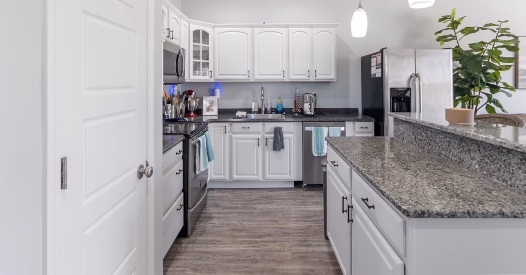 A white kitchen with dark countertops, stainless steel appliances, hanging lights, and a potted plant on a bar.
