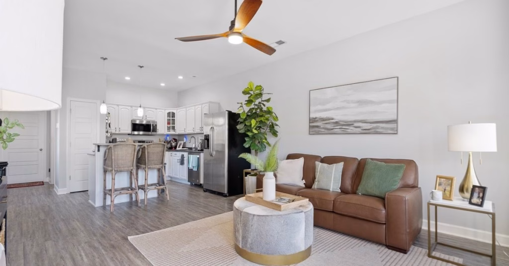 An open ADU living space features a brown sofa and round ottoman beside a white kitchen with island seating.