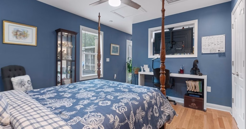 A blue bedroom contains a floral four-poster bed, a wall-mounted TV, a desk, a ceiling fan, and hardwood floors.