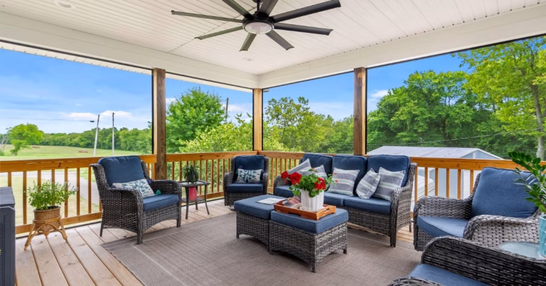 A covered patio contains blue wicker seating, a coffee table, potted plants, and a ceiling fan above.
