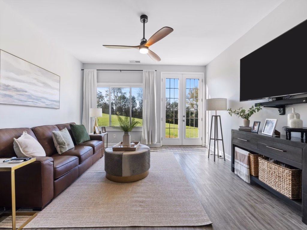 A bright living room features a brown sofa, a large wall-mounted TV, French doors, and wide backyard views.
