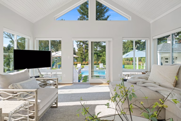 A bright room addition features a vaulted ceiling, large windows, a sofa, chairs, and doors overlooking a backyard pool.