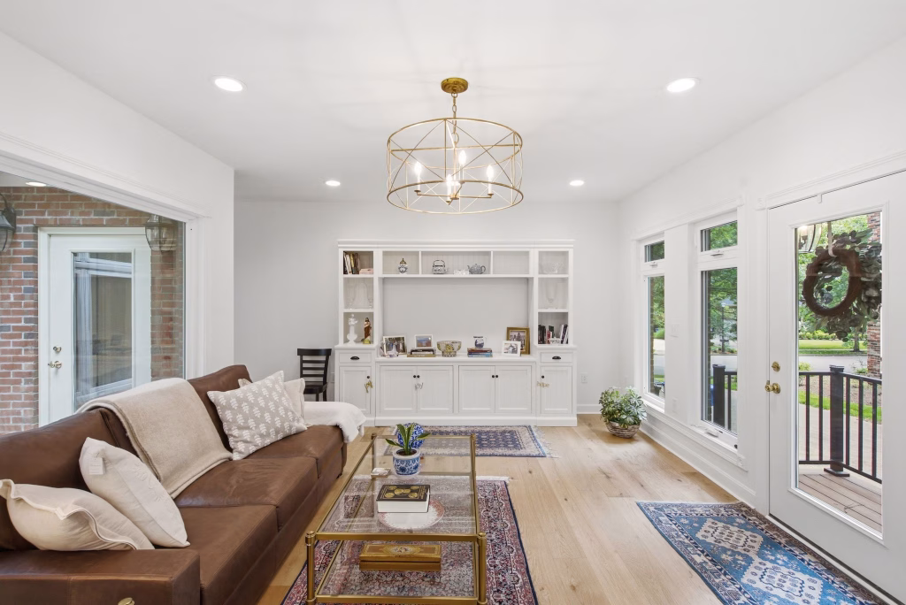 A bright sitting room features a brown sofa, built-in white shelving, large windows, and a glass door leading outside.