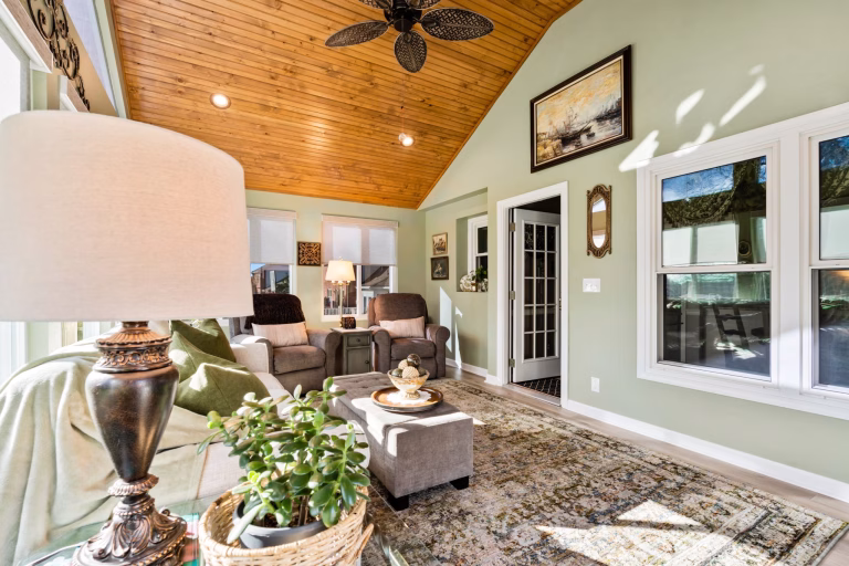 A vaulted sunroom features a warm wood ceiling, large windows, a sofa, two armchairs, and a patterned area rug.