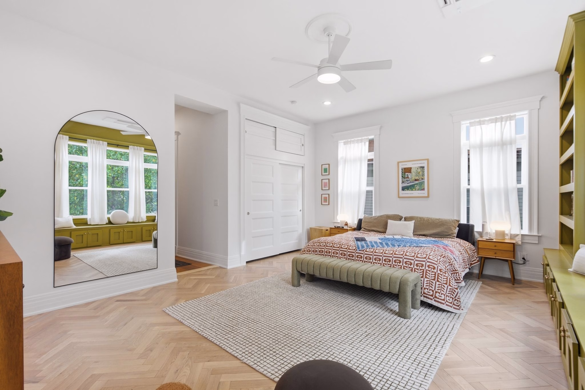 A bright bedroom features a large bed, tall windows, a ceiling fan, built-in shelving, and a wide mirror on the wall.