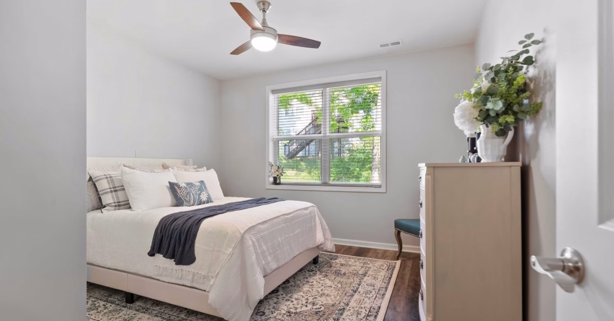 A bright bedroom contains a white bed, a wood dresser, a large window, and a ceiling fan above a patterned rug.