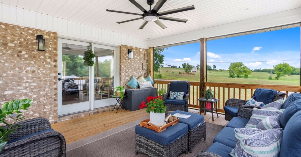 A covered patio containing blue wicker seating, potted plants, and a ceiling fan overlooks a wide green field.