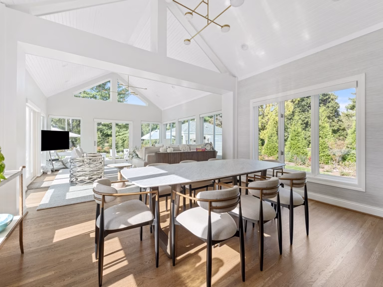 A long dining table fills a bright open room with a white vaulted ceiling, large windows, and a connected sitting area.