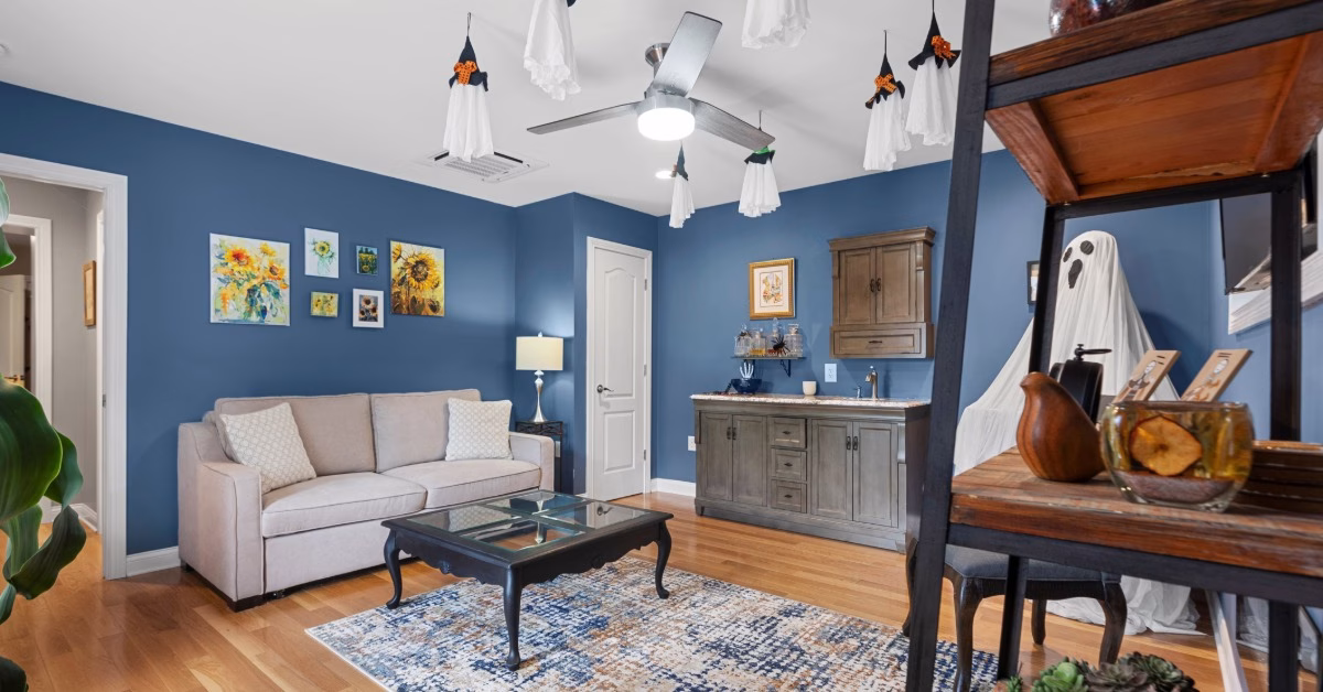 A beige sofa sits in a blue living room with a wet bar, hardwood floors, a coffee table, and hanging ghost decorations.