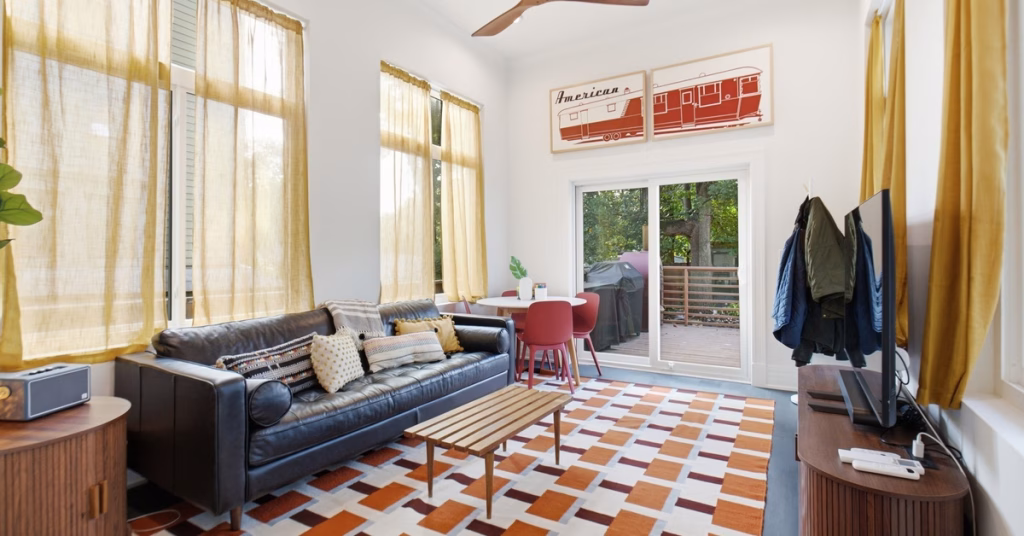 A beige room contains a gray sofa, floral chair, wide windows, a ceiling fan, and views of a green backyard.
